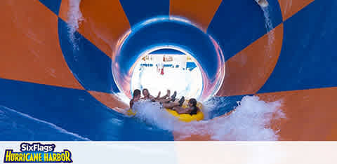 Image shows a group of people in a yellow raft sliding down a blue and orange water slide at Six Flags Hurricane Harbor. There is visible excitement as they are surrounded by splashes of water, with bright daylight waiting at the end of the tunnel. The park's logo is displayed at the bottom.