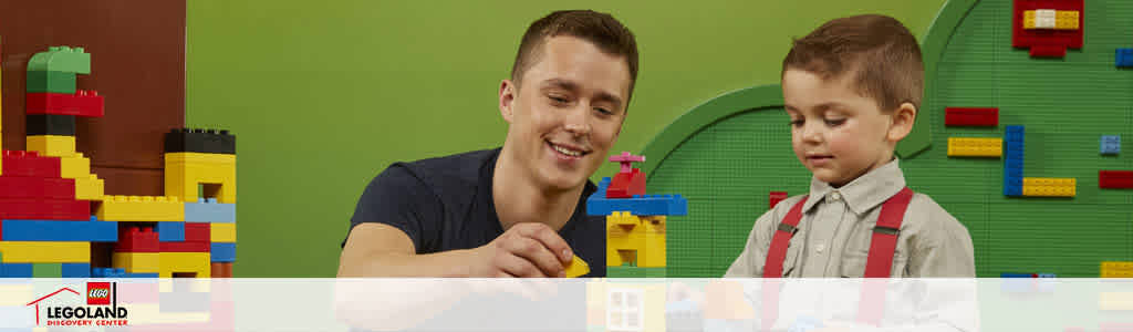Image shows an adult and a child engaging with colorful building blocks at LEGOLAND Discovery Center. They seem joyful and focused on constructing a structure. A vibrant green backdrop with built-in LEGO designs enhances the playful atmosphere.