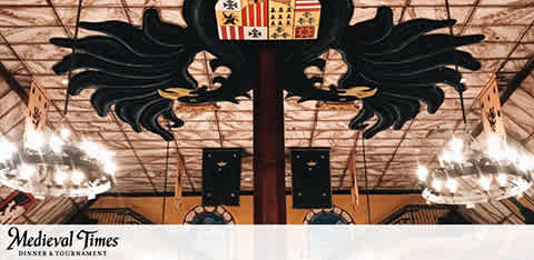 The image shows the intricate interior of a themed dining area, evoking a medieval hall. A decorative shield with a coat of arms is centered below an elaborate black sculpture adorned with feathers, resembling a crest or a pair of wings. Warm lighting is cast from chandeliers above, and the space hints at the anticipation of a theatrical dinner show. The words 'Medieval Times Dinner & Tournament' are displayed at the bottom, indicating the venue's name.