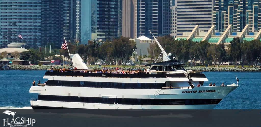 This image shows a large, white multi-deck cruise ship named 'Spirit of San Diego' floating on the water with a clear view of its port side. The ship, adorned with the 'Flagship Cruises & Events' branding, is filled with passengers on its multiple open-air decks. Many passengers are visible enjoying the sunny weather and the view. In the background, the city skyline of high-rise buildings lines the waterfront, and a few trees from a nearby park can be seen. The American flag flies proudly atop the rear of the ship.

For an unforgettable experience on the waves and to make your trip even more enjoyable, FunEx.com offers unbeatable discounts on tickets, ensuring you get the lowest prices for your maritime adventure.