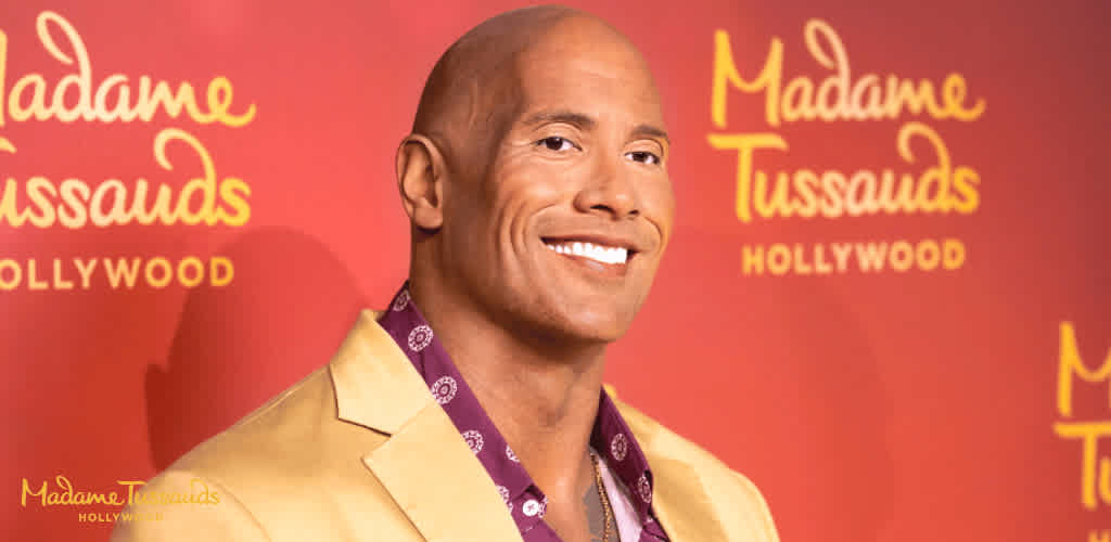 A man in a beige blazer and purple shirt smiling against a red Madame Tussauds Hollywood background.