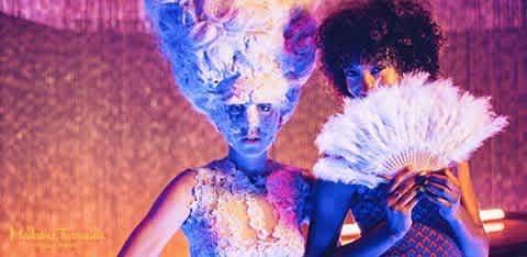 Two people with vibrant hair and elaborate costumes pose in dramatic lighting with one holding a large white fan.