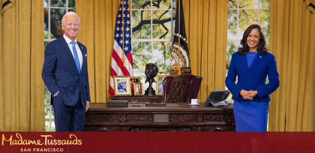 This image features two life-sized, realistic wax figures that are dressed professionally and are standing in a room that strongly resembles the Oval Office. On the left, the male figure is wearing a dark blue suit, white shirt, and light blue tie. He is smiling broadly and standing with one hand at his side and the other slightly extended. On the right, the female figure is dressed in a blue suit with a skirt and is standing with her arms crossed, also smiling. Behind them, an American flag and a flag with an eagle atop a crest can be seen alongside heavy gold drapes that frame a window showing trees and sunlight. The desk between them has several framed photographs, a telephone, and a bronze bust. The foreground includes a red banner with the text "Madame Tussauds San Francisco" inscribed in gold lettering that informs the viewer of the location where these wax figures are displayed.

At FunEx.com, we offer the thrill of experiencing such lifelike figures at a fraction of the cost, ensuring you enjoy the excitement without straining your wallet. Discover our exclusive deals for discounted tickets and savings on your next fun-filled outing.