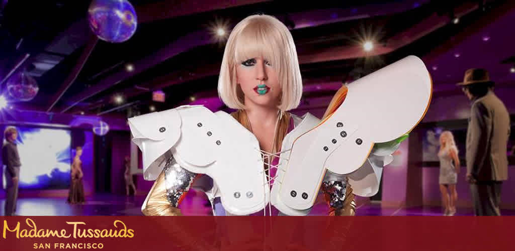 This image features a vibrant scene from Madame Tussauds San Francisco. In the foreground, there's a life-sized, photorealistic wax figure of a pop star with short, straight blonde hair and striking makeup, including bright blue lipstick. The figure is dressed in a silver, glittery outfit paired with oversized, wing-like white shoulder adornments that resemble a fusion of futuristic fashion and angel wings. Behind the figure, the scene is set in a room bathed in purple lighting with disco balls suspended from the ceiling, adding a lively and glamorous atmosphere. Visitors are present in the background, viewing the exhibitions. Notably, the room hosts additional wax figures, enhancing the interactive and celebratory feel of the venue. At FunEx.com, you can anticipate not only the excitement of renowned attractions but also the benefit of impressive savings, as we offer the lowest prices and exclusive discounts on tickets to enrich your experiences.