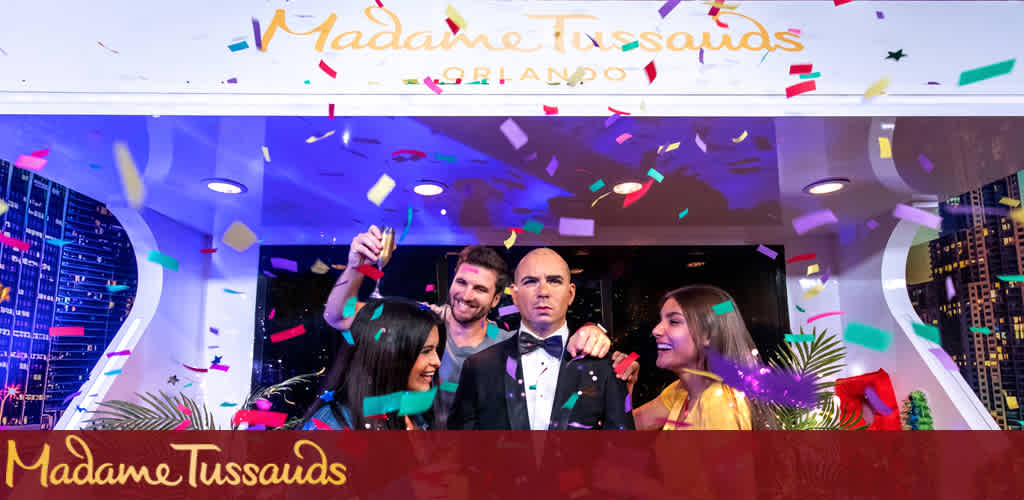 This image features a joyful moment at Madame Tussauds Orlando with a colorful confetti celebration. Two women and a man, presumably visitors, are seen smiling and taking a selfie with a wax figure resembling a well-known celebrity dressed in a black suit and tie. They are standing behind a white counter with the iconic Madame Tussauds signage overhead, embellished with multicolored confetti strewn in the air, which adds a dynamic and festive atmosphere to the scene. The backdrop illustrates a cityscape at night, enhancing the feeling of a vibrant nightlife event. Remember to book your experience through FunEx.com, where we promise sensational savings and the lowest prices on tickets for your next memorable outing.