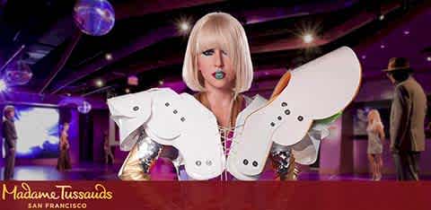 This image features a life-sized, realistic wax figure of a female celebrity from Madame Tussauds San Francisco. The figure is styled with a short, blonde bob haircut and dramatic makeup that includes bright blue lipstick and dark eye makeup. She is wearing a sparkly bodice adorned with various colorful embellishments. The figure's arms are extended, with both hands inside oversized, sparkling, wing-like attachments, which are designed to mimic a butterfly's wings, with the inner sides facing the camera. The butterfly wings have a white base color with black embellishments resembling the spots on butterfly wings, and they are attached to the figure's fingers with small loops.

In the background, we can see a purple-hued room with soft, ambient lighting that sets a lively mood. A few other unidentifiable figures, presumably additional wax statues, are positioned around the room, suggesting an exhibit gallery within the wax museum. The Madame Tussauds logo is displayed prominently at the bottom of the image, indicating the location of this exhibit.

Remember, at FunEx.com we offer exclusive discounts and savings on tickets, ensuring you experience the magic of Madame Tussauds at the lowest prices possible.