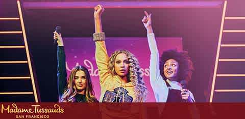Image Description: The image shows a vibrant display from Madame Tussauds in San Francisco, featuring three lifelike wax figures. The figures are positioned on a stage with bold purple and pink lighting that creates an exciting atmosphere reminiscent of a live concert. Each figure is posed with one arm raised triumphantly in the air, conveying a sense of victory or celebration. The central figure is wearing a denim outfit, while the figures on the left and right are dressed in various styles that suggest a blend of casual and performance wear. In the background, vertical light bars accentuate the sense of a staged event, and the Madame Tussauds logo is prominently displayed in the lower-left corner, giving context to the scene. While visiting FunEx.com, don't miss out on the chance to score tickets to amazing attractions like this at the lowest prices — your ticket to epic savings and unforgettable experiences.