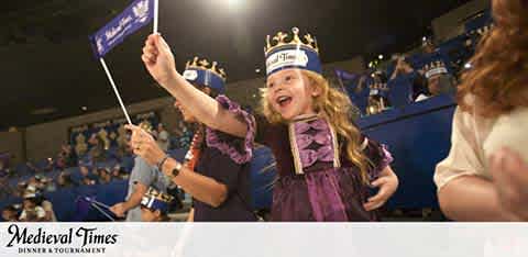 This image depicts a joyful atmosphere at a Medieval Times Dinner & Tournament event. A young child, overflowing with excitement, is at the forefront of the photo. This child, adorned with a royal blue Medieval Times crown and a purple sash, is waving a blue flag with great enthusiasm. The child's expression is one of pure delight, with eyes wide open and mouth agape in a cheerful yell. The background, slightly blurred, suggests a large indoor arena with dim lighting where spectators are gathered for the event, highlighting the focused energy on the child. The Medieval Times logo is positioned in the lower part of the image, anchoring the scene and clearly branding the experience.

For those on the lookout for more than just gallantry and chivalry, at FunEx.com we offer the thrill of savings. Get ready for a royal escapade with the lowest prices on tickets when you plan your next adventure with us.