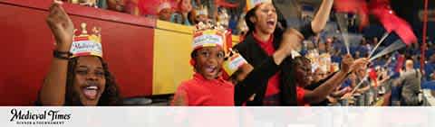 This image showcases a vibrant scene at a Medieval Times event, capturing the enthusiasm and joy of the audience. On the left, a young girl with a bright smile is holding a red flag, her eyes lit up with excitement. Next to her, another child is clapping, wearing a paper crown that is also donned by many audience members, suggesting a shared experience of medieval pageantry. To the right, a woman is seen cheering passionately, with a red flag in one hand. Everyone is focused on the action happening out of view, presumably a jousting match or another form of medieval entertainment. The crowd is seated in a stadium-style arena with red seating, reinforcing the theme of the event. The words "Medieval Times" are displayed in the lower right corner, indicating the brand or venue associated with the experience. For visitors planning to engage in this lively and historical entertainment, FunEx.com offers not just a memorable outing but also the promise of unbeatable savings and the lowest prices on tickets.