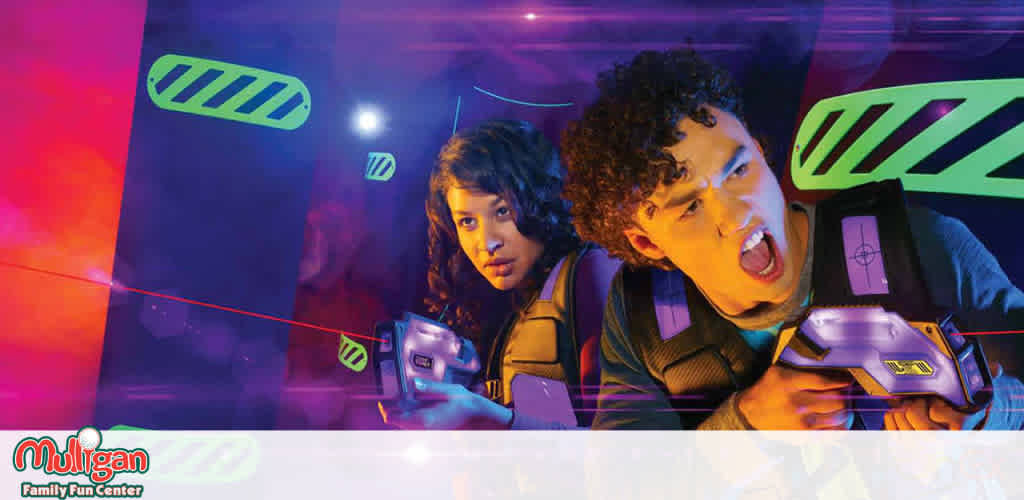 This image features two young individuals engaged in a laser tag game at the Mulligan Family Fun Center. They are positioned side-by-side, amidst a vibrantly lit arena that glows with purple and pink hues. Both are wearing black vests adorned with neon lights and are geared with futuristic-looking laser guns, one in each hand, pointing directly ahead. Red laser beams streak across the view, adding to the dynamic and action-packed atmosphere. Neon green lights in the shape of elongated hexagons adorn the walls, contributing to the high-energy, gaming environment. At the bottom left, the Mulligan Family Fun Center logo is prominently displayed, signaling a place of entertainment and group activities.

Remember, at FunEx.com, you can enjoy the thrill of adventure at Mulligan Family Fun Center for less, with exclusive discounts, the lowest prices, and a seamless ticket purchasing experience!