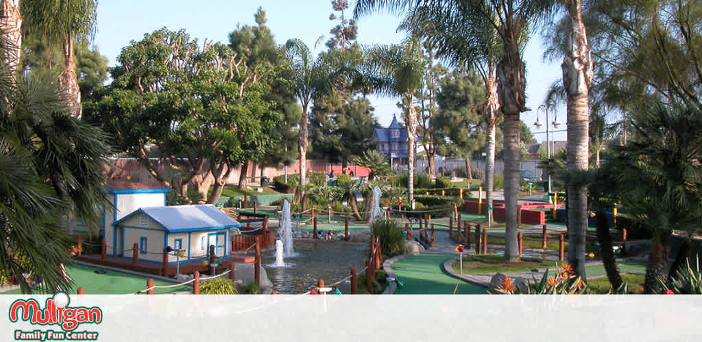 This image features a vibrant outdoor scene at Mulligan Family Fun Center, showcasing a miniature golf course with a beautifully landscaped setting. In the foreground, palms and other trees frame the view, partially obscuring details and adding depth. The main focus is a central miniature golf area with bright green artificial turf and red-trimmed borders, where holes are interspersed with small water features, including a water fountain. A quaint, blue-roofed building resembling a cottage serves as a focal point within the greenery and the golf area. In the distance, more green foliage and trees can be seen under a clear sky, suggesting a tranquil, family-friendly entertainment environment.

At FunEx.com, we're committed to providing the joy of miniature golf with the added bonus of savings, offering the lowest prices and great discounts on tickets to ensure your visit to Mulligan Family Fun Center is both delightful and affordable.