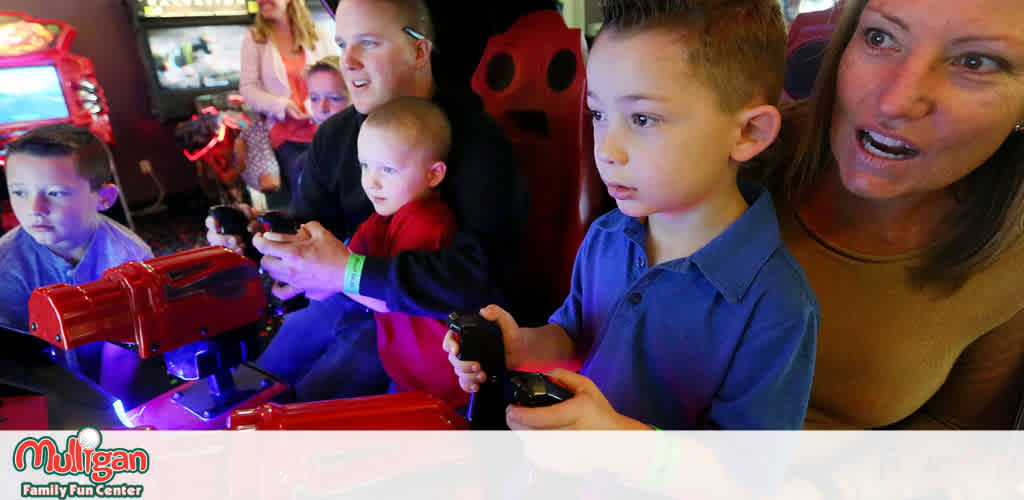 Image Description:
The photograph captures a moment at Mulligan Family Fun Center, showcasing a joyful arcade experience. In the foreground, right of center, a young boy in a blue shirt is focused intently on a video game, his hands grasping a small black controller. To the left, a younger child in a red shirt, also holding a controller, sits beside a smiling adult man, who is engaged in playing the game with the child. All three are intently watching the screen in front of them, absorbed in the excitement of the game. Slightly behind this group, to the left, another young boy in a dark blue shirt is visible, also concentrating on a game controller. In the right background, there is a woman in a brown top, enthusiastically cheering on the players, with her attention directed towards the screen and action. Faintly behind her, a girl with her hair in braids is partially visible, observing the gaming fun. The ambiance is lively and filled with vibrant colors of the arcade machines, creating a sense of fun and family entertainment.

For unbeatable savings on your next adventure, visit FunEx.com where you can find the lowest prices on tickets to a wide array of attractions, including family fun centers just like this one!