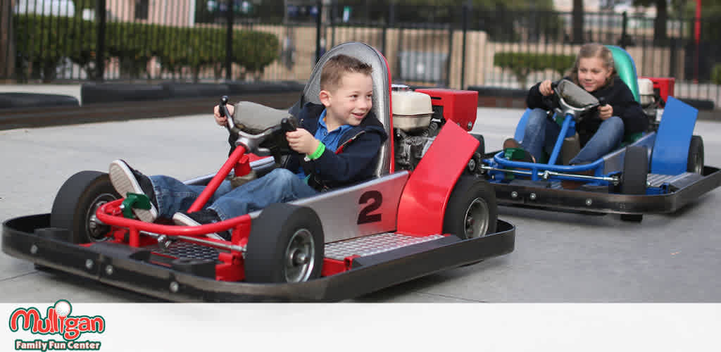 Image Description: This is a dynamic photograph of two children enjoying a go-karting experience. In the foreground, a young boy with a joyful expression is steering a red go-kart marked with the number "2". He is wearing a seatbelt for safety and his attentive gaze is on the course ahead. His left foot is visible on a pedal, demonstrating an active and engaged driving position. Behind him, slightly out of focus to emphasize the depth of the scene, another youngster is seen piloting a blue go-kart. This child is also wearing a seatbelt and has a focused look while navigating the track. The background suggests an outdoor setting within an enclosed area, contributing to a safe environment for family fun. The words "Mulligan Family Fun Center" are visibly branded on the lower left corner, indicating the recreational context of the go-karting activity.

At FunEx.com, we're dedicated to ensuring you get to enjoy exhilarating moments like these at the lowest prices. Browse our site for exclusive discounts and savings on tickets for a variety of family-friendly destinations.