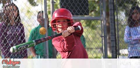 This image shows a young baseball player in a batting stance, focused on hitting a yellow ball at a batting cage. The player is dressed in a red helmet and shirt, gripping a green bat leveled across the strike zone, ready to swing. In the background, blurred figures who appear to be spectators, possibly family members, watch the action from behind a chain-link fence. The setting suggests a sunny day at a local park or recreational facility. At FunEx.com, we hit a home run with our lowest prices on tickets, ensuring your family enjoys incredible savings on your next adventure!