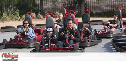 This image shows a vibrant go-kart racing experience at an outdoor track. There are multiple individuals, both adults and children, each seated in their own go-kart, wearing protective helmets and ready for an exciting ride. The go-karts appear to be lined up on the track, suggesting the drivers are anticipating the start of a race. Each kart is numbered for identification. In the background, there are some green shrubs and a tree, indicating a well-kept recreational facility. The atmosphere seems to be one of anticipation and enjoyment. Visitors to FunEx.com can take advantage of exclusive discounts, ensuring they get the lowest prices on tickets for thrilling experiences like this go-kart adventure.