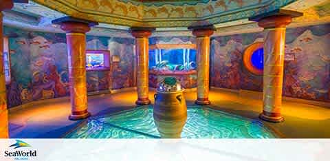 Image Description: An indoor SeaWorld attraction featuring a vibrant, undersea-themed room. Decorative columns support a domed ceiling with elaborate murals depicting marine life. Circular windows offer a glimpse into aquatic habitats, while the center of the room displays a prominent, ornate vase. The floor mimics an oceanic pattern, enhancing the subaquatic ambiance. The SeaWorld logo is visible in the corner of the image.