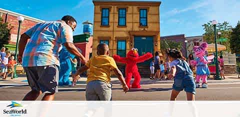 Image features a bright outdoor scene at a theme park with visitors engaging with costumed characters. Children excitedly approach a red, furry character in the center, while a man in a blue shirt gestures towards it. In the background, other characters and people are visible, and a blue sky adds to the lively atmosphere. A  SeaWorld  logo is at the bottom.