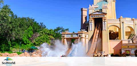 Image shows a water ride at SeaWorld with a log flume splashing down into the water, creating a large spray. The surrounding setting includes a sunny sky, a palm-tree-lined landscape, and a pastel-hued building facade typical of the park's aesthetic.