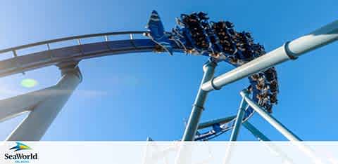 Image shows a roller coaster with riders inverted at the peak of a loop. The track is a pale blue set against a clear sky. The coaster is filled with thrilled riders, legs dangling in the air. SeaWorld logo is visible at the bottom. Excitement under the sun.