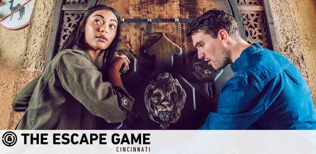 Image displays two individuals engaged in a puzzle at The Escape Game Cincinnati. A woman looks outward while reaching into a wall-mounted feature, and a man concentrates on adjusting something within it. Both appear immersed in solving the challenge, hinting at the fun, interactive experience offered by the escape room. The logo of The Escape Game is visible in the corner.