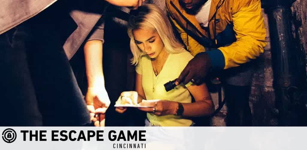 Image shows a group of people engaged in an activity at The Escape Game Cincinnati. A woman in the foreground examines a clue illuminated by a flashlight, which a man holds over her shoulder. Other participants look on, indicating a collaborative effort in a dimly lit, adventure-themed setting.