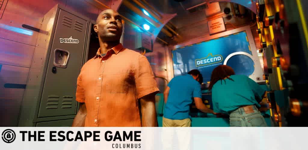 Image of a man standing in the foreground, looking off to the side with an intrigued expression. He's inside a room themed like a submarine with ambient lighting. In the background, three individuals in blue shirts are engaged with the equipment, possibly solving puzzles. The text,  THE ESCAPE GAME COLUMBUS,  is displayed, and a logo with the word  DESCEND  is visible in multiple places.