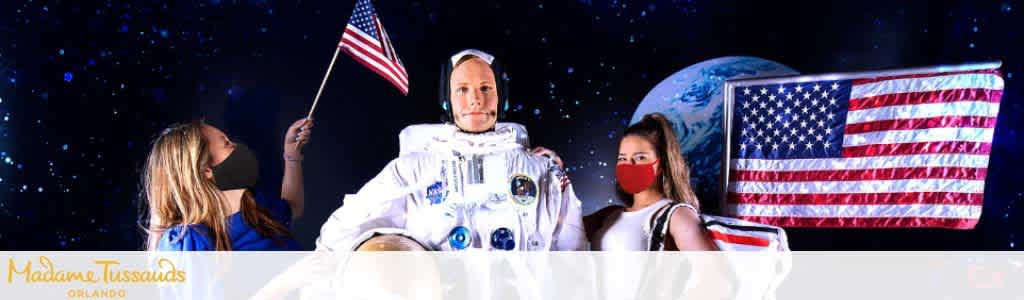 Two individuals with masks near a wax astronaut holding an American flag, with a starry backdrop.