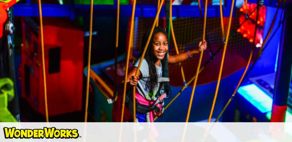 A girl in a harness smiles while navigating a colorful indoor ropes course.