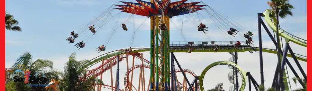 Swing ride in motion with people flying above a theme park with roller coasters and palm trees.