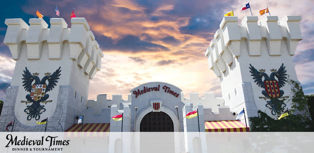 The image is a promotional photo of a Medieval Times castle facade during dusk. It showcases a large, castle-like building with white walls and two stone towers adorned with banners and colorful flags. A dramatic sky with a sunset forms the backdrop. The words Medieval Times Dinner & Tournament are displayed prominently. A central crest features an eagle on the entrance.