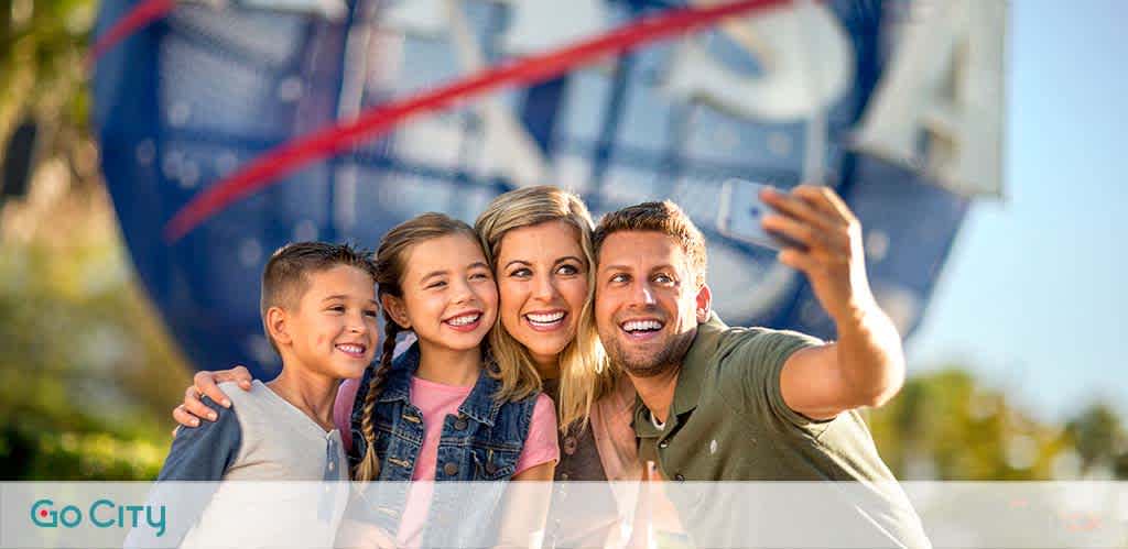 Family of four takes a selfie, smiling outdoors on a sunny day, with a Go City banner in the background.