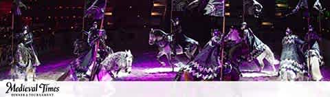 Image shows a dramatic medieval-themed performance with horse riders in ornate costumes. Cavaliers in purple and black attire on horseback, demonstrating skillful riding in an arena illuminated by spotlight. The Medieval Times logo is visible, hinting at an engaging, historical dinner show experience.