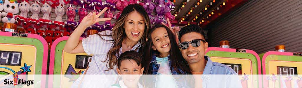 A joyful family of four is shown at an amusement park, posing in front of colorful game stalls with plush prizes visible in the background. The mother gives a peace sign while embracing a boy, the father smiles widely with sunglasses on, and a girl leans on his shoulder, all displaying happiness.