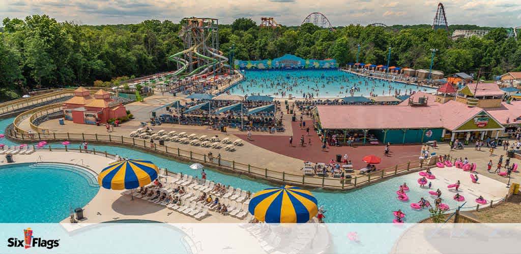 This image shows an expansive outdoor water park on a sunny day with large pools and a crowd enjoying the water. Lounge chairs line the poolside under colorful umbrellas. Water slides tower in the background while a wave pool is filled with swimmers. Flamingo-shaped floaties add a playful touch near a sandy area. Trees and roller coasters are visible in the distance, indicating the park is part of a larger amusement facility. The sky is partly cloudy, suggesting pleasant weather for water activities.