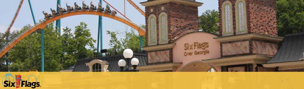 Image shows the entrance facade of Six Flags over Georgia with a roller coaster filled with people in the background. The coaster is ascending on tan tracks against a clear blue sky. Below is a yellow banner featuring the Six Flags logo.