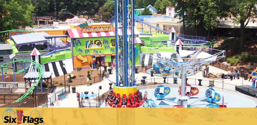 Colorful amusement park section with a blue sky background. A prominent 'FUNHOUSE' sign in multicolored letters is seen above the entrance to an attraction. Various rides including a green water slide, a spinning ride with blue seats, and a drop tower with red seats are visible. Guests are seen walking and enjoying the park. The Six Flags logo is in the bottom left corner.