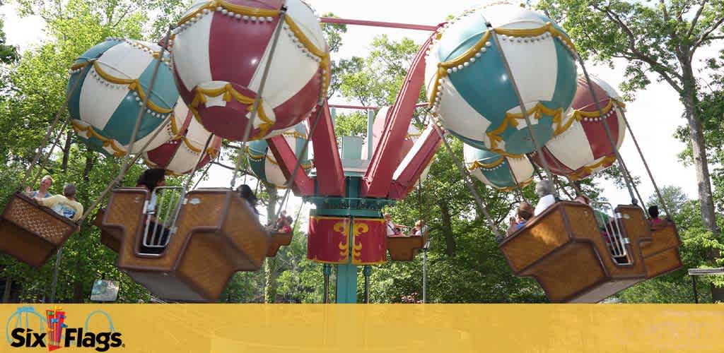 Ride at Six Flags amusement park featuring colorful hot-air balloon replicas. Each balloon has a basket carrying visitors, suspended against a backdrop of green trees under a clear sky. The balloons are adorned in white, red, and blue with gold accents. The Six Flags logo is visible, indicating the location.