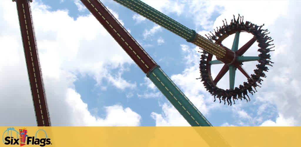 Image shows a thrill ride at a Six Flags amusement park on a sunny day with a blue sky and clouds. A towering vertical green track holds a large rotating wheel with seats facing outward, filled with riders experiencing the excitement. The Six Flags logo is visible on the bottom left.