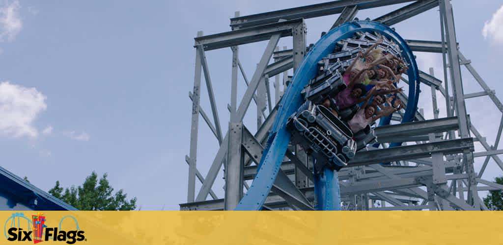 Image of a roller coaster at Six Flags amusement park with excited riders upside down at the peak of a loop. The structure is a mix of white and blue steel against a clear sky. The Six Flags logo is visible in the upper corner.