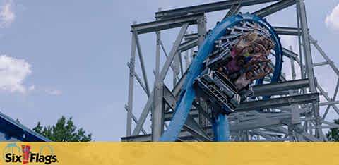 Image of a blue roller coaster with excited riders descending a steep track. A metal structure supports the ride against a clear blue sky. In the foreground, the Six Flags logo is prominently displayed above a yellow stripe.