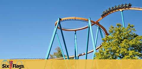 Image shows a section of a roller coaster with a blue track and brown supports against a clear sky. In the foreground, a yellow wall partially obscures the view. The logo of Six Flags is visible, indicating the location is one of their amusement parks.