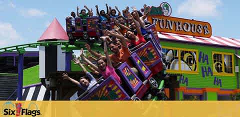 Thrilled riders on a colorful roller coaster descent at Six Flags amusement park. Arms raised in excitement against a backdrop of a playful funhouse with bold 'HA HA' decals and vibrant patterns. Bright, sunny day enhances the exhilarating atmosphere.