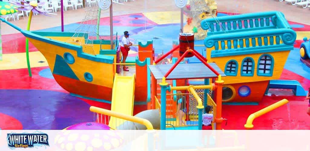 Colorful children's water play area with a large yellow and blue ship-like structure featuring slides and water cannons. A smaller boat and playhouse with climbing nets complete the playful scene. The ground is splashed with vibrant blues, reds, and yellows.
