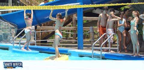 Image features excited visitors at a water park about to enjoy a slide. Two people are on the slides, while others wait their turn. The environment is sunny, with a bright blue slide structure. The watermark 'WHITE WATER' indicates the park name.