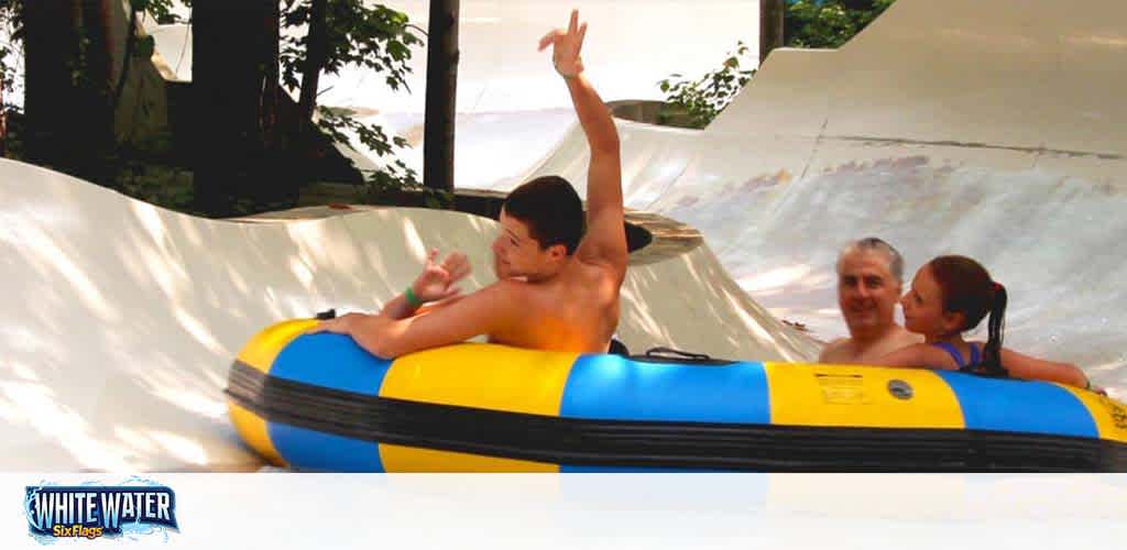 Three individuals are riding a yellow and blue raft down a water slide, surrounded by foliage. The person in front raises their arms in excitement, while the two behind show enjoyment. The Six Flags White Water logo is visible in the corner.