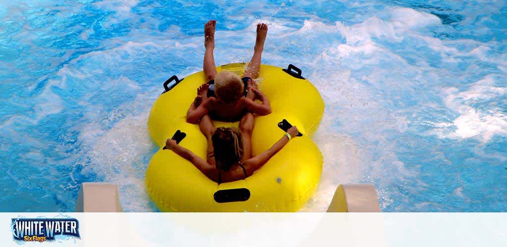 Two individuals are enjoying a water ride in a yellow double tube. The person at the back raises a hand in excitement while moving down a blue water slide with splashes around them. The logo in the corner shows 'White Water Six Flags,' indicating the location is a water amusement park.