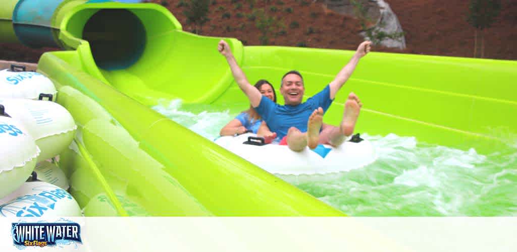 Image shows two people, a man and a woman, enjoying a water slide ride in a circular raft. The man is in the front, arms raised in excitement, while the woman is behind him, both smiling. The slide is bright green and they are emerging from a tunnel into an open area with water splashing around them. The logo of White Water Six Flags is visible on the side, indicating the location is an amusement park. The setting seems to be outdoors with daylight.