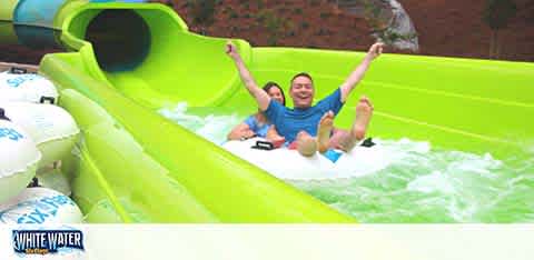 Image of a water slide with bright green flume. Two people are enjoying a ride in a blue raft with arms raised in excitement. There's splashing water around them indicating motion. The slide exit appears in the foreground and the company logo White Water is visible at the bottom.