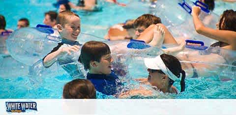 The image shows a lively scene at a water park. Children and adults float in a bright blue pool, with some on inflatable rings. Splashes of water and the joy of swimming outdoors are evident. A logo at the bottom reads 'WHITE WATER'.