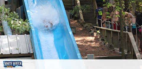 The image displays an outdoor water slide at a water park, with a person in a splash of water at the end of the slide. Onlookers stand by the sides, partially obscured by trees and foliage. The water slide is branded with 'WHITE WATER.' The setting appears to be in a forested area on a sunny day.